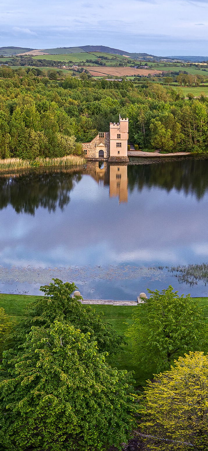 An aerial view of Oakfield Park castle and lake in daylight during summer. The castle is reflected in calm water, surrounded by trees with lush green foliage.