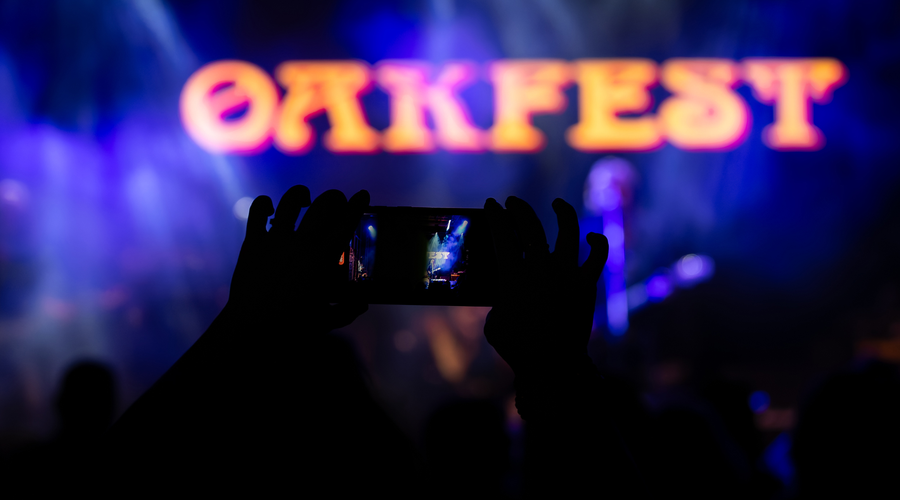 Oakfest 2026 header image showing a person in the audience using a smartphone to take a photo of the stage and Oakfest logo on the backdrop